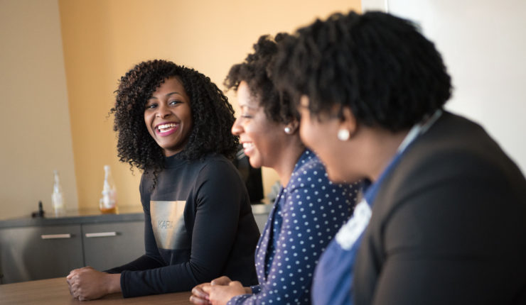 A group of 3 diverse women sat at a board table, one of whom is laughing