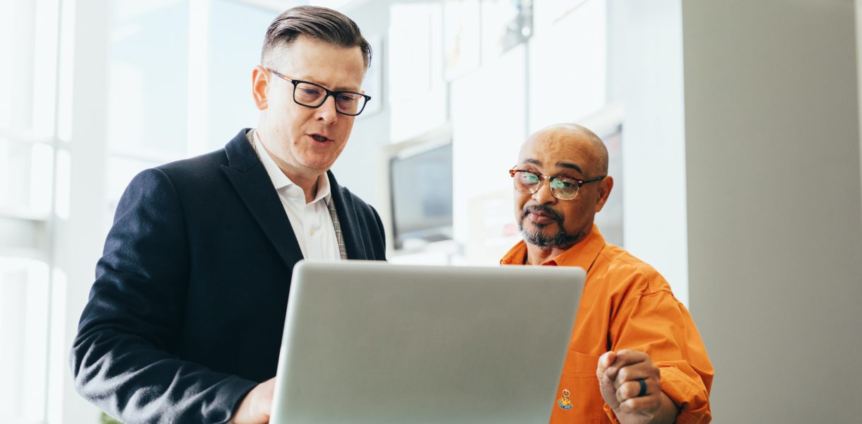 two men talking at table looking at a laptop